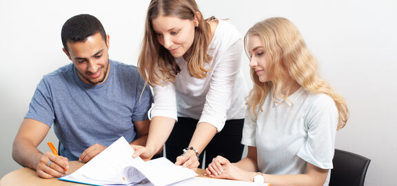 Ein junger Mann und zwei junge Frauen blicken bei Tisch in eine geöffnete Mappe